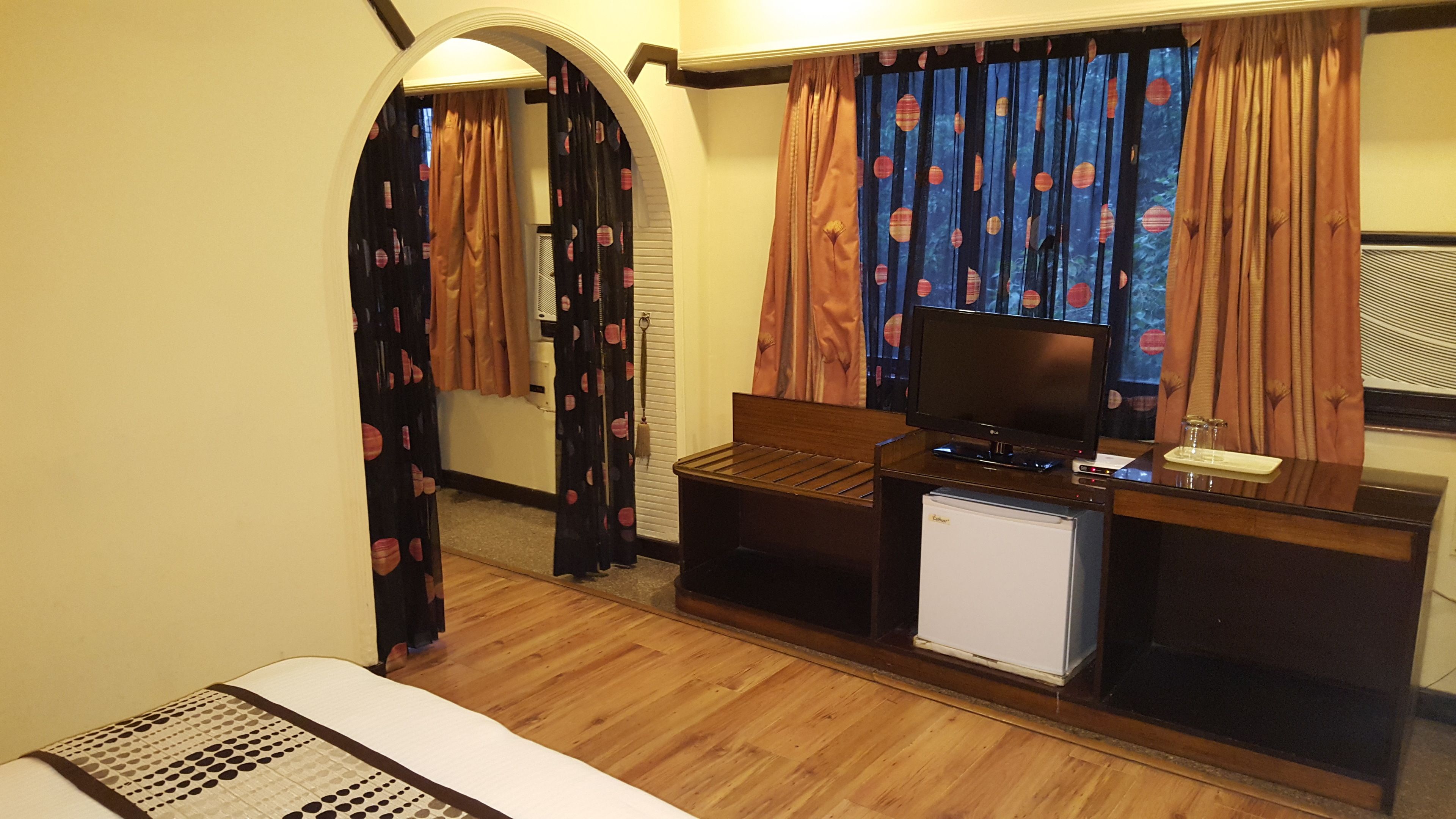 A compact hotel room with a bed, a small desk with a television, a mini-fridge, and a window with curtains.