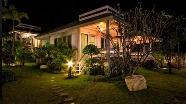 Una casa moderna de dos pisos con una entrada principal bien iluminada, una escalera visible y un jardín ajardinado con plantas tropicales.