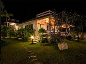 Una casa moderna de dos pisos con una entrada principal bien iluminada, una escalera visible y un jardín ajardinado con plantas tropicales.