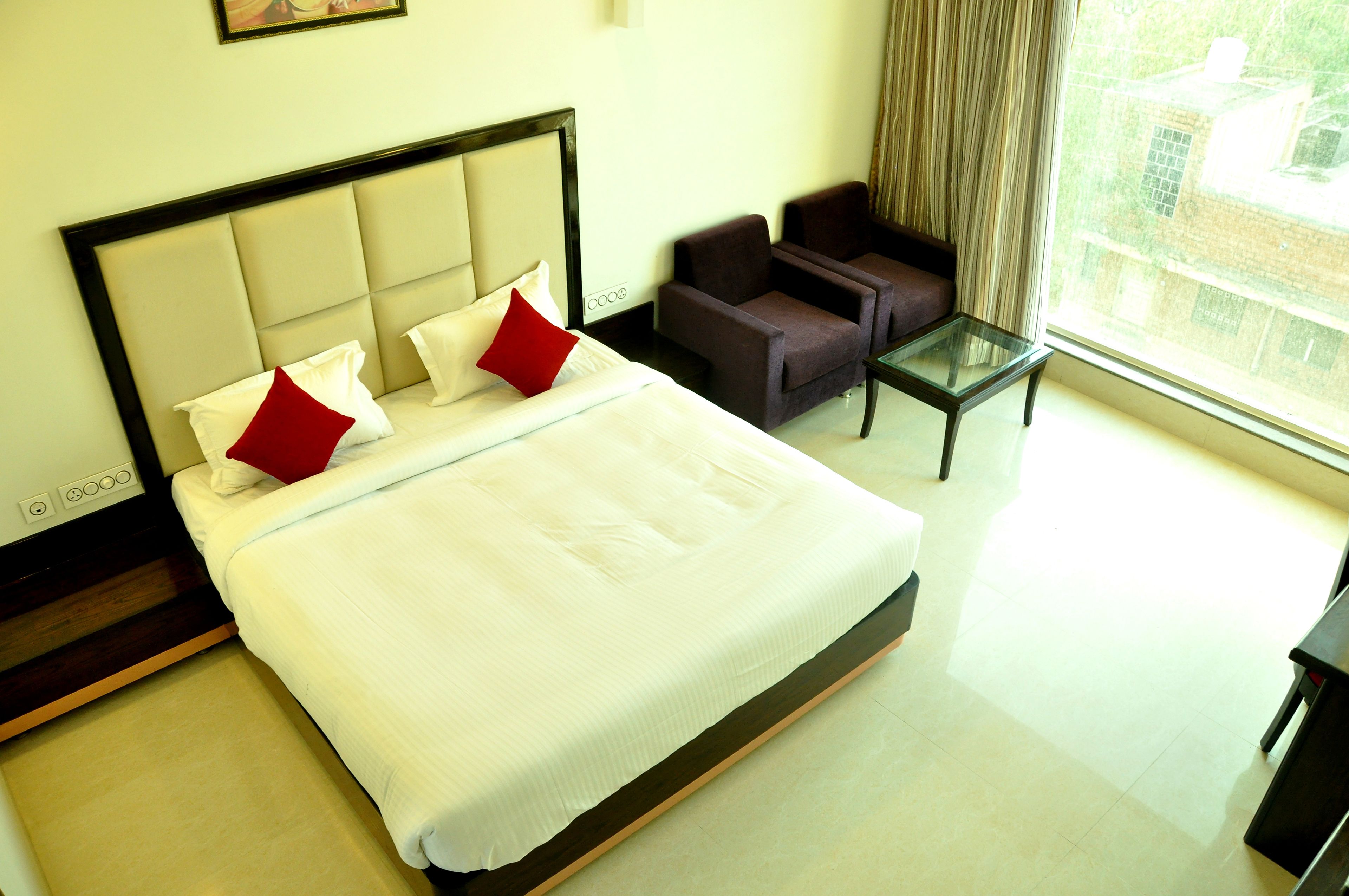 Habitación de hotel con una cama grande, dos sillones, una mesita y un ventanal con vistas a edificios.