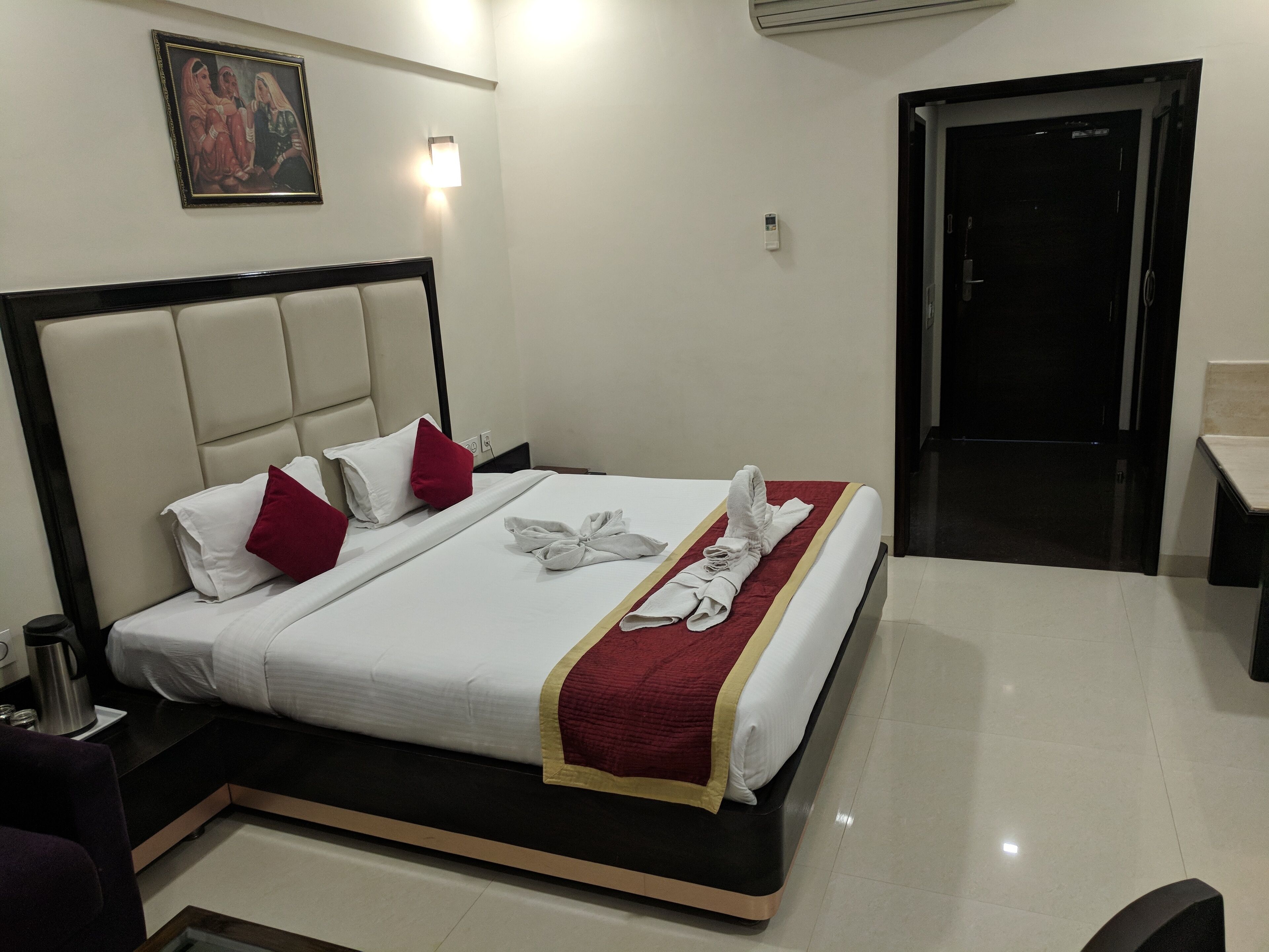 Habitación de hotel con cama, almohadas, toalla, un cuadro, mesita de noche, cafetera y puerta.