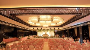 Un salón de banquetes con filas de sillas, un escenario con adornos florales y una gran lámpara de araña.