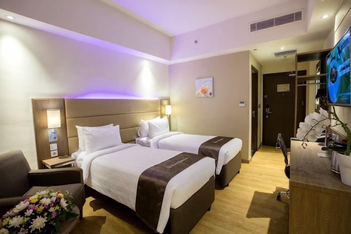 A hotel room with two beds, a desk, a chair, a TV, and a reception desk.