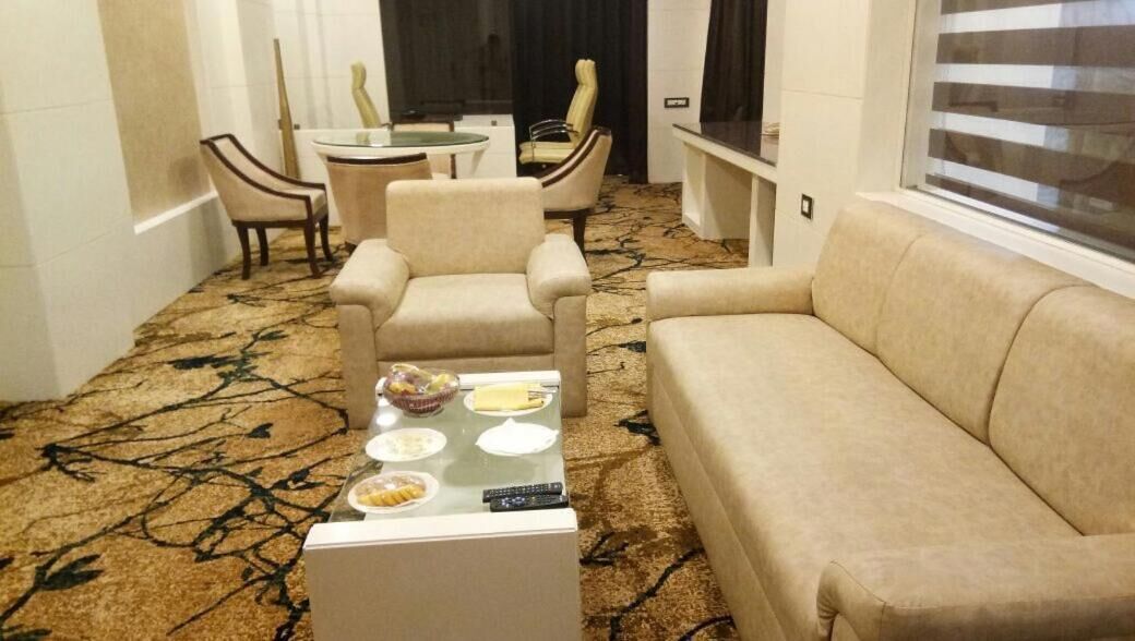 A living room with a beige sofa, two armchairs, a coffee table with plates and a remote control, and a patterned carpet.