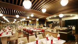 Um restaurante espaçoso com várias mesas de jantar, cada uma com toalhas brancas, guardanapos vermelhos e talheres. O teto apresenta um design de madeira com luminárias circulares.