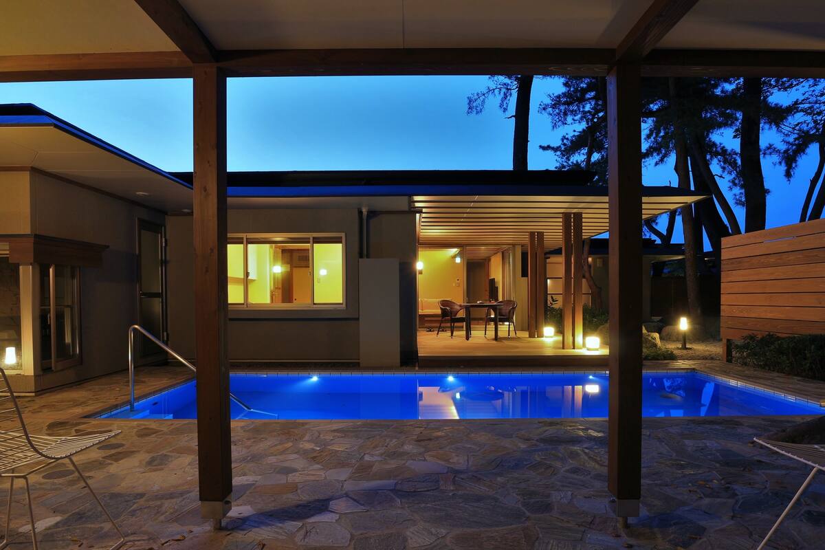 A modern house with a swimming pool, stone flooring, and a covered patio area with a table and chairs.