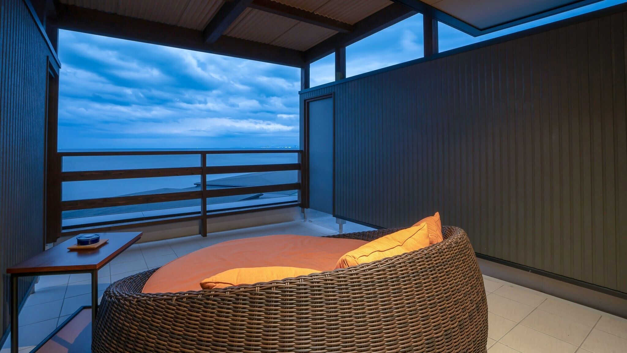 Un balcón con una silla de mimbre, una mesita y vista al océano.