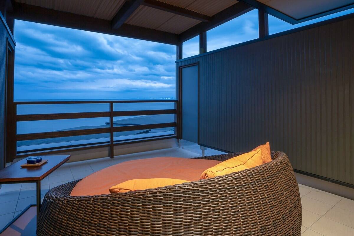 A balcony with a wicker chair, a small table, and a view of the ocean.