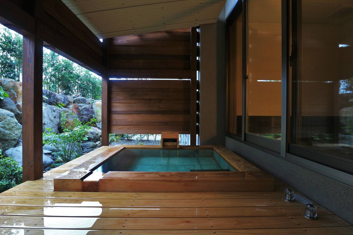 A wooden deck leading to a hot spring bath area with a view of rocks and greenery.