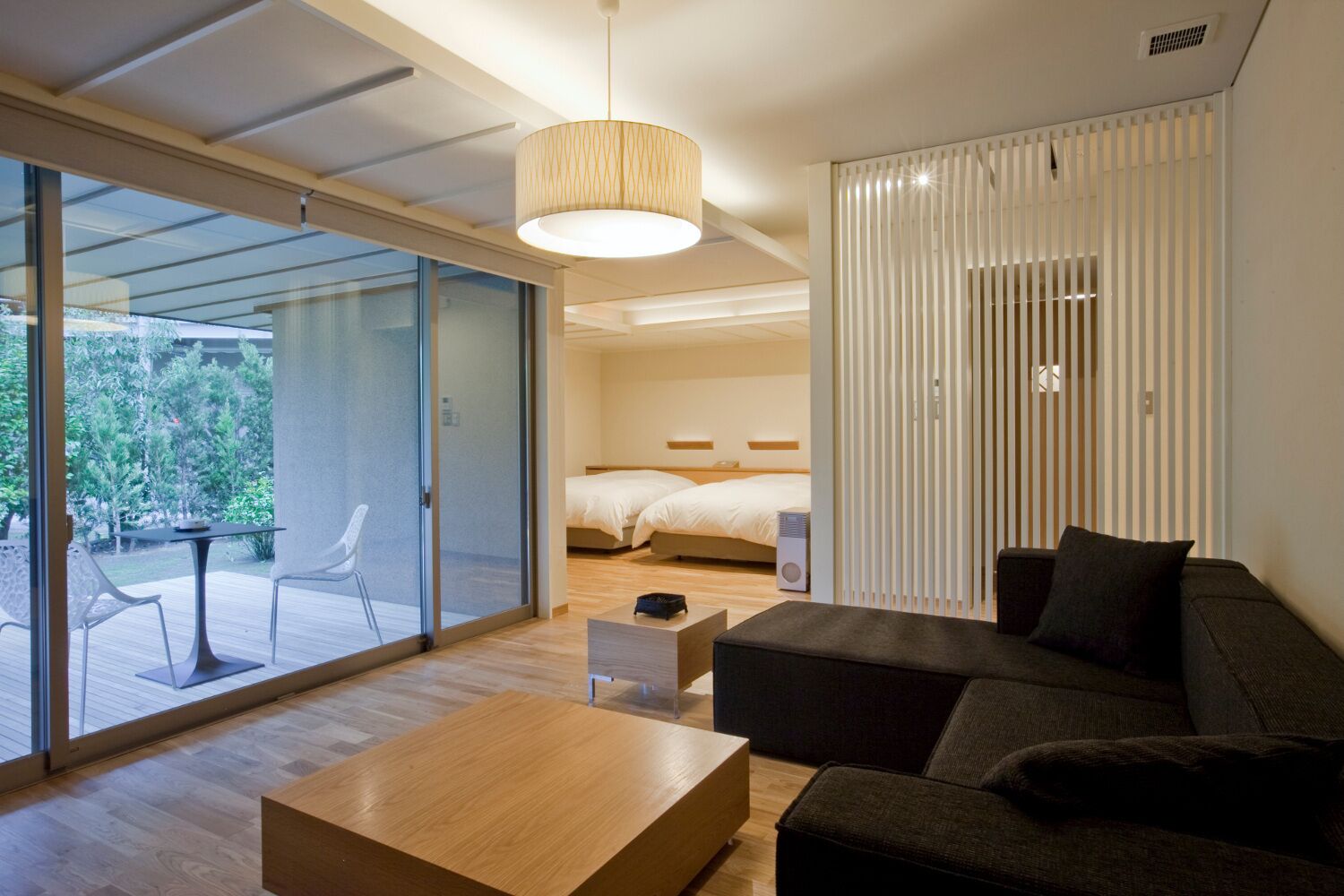 Ett modernt hotellrum med en skjutdörr i glas som leder ut till en uteplats, en säng med sänggavel, en soffa, ett soffbord i trä och en hänglampa.