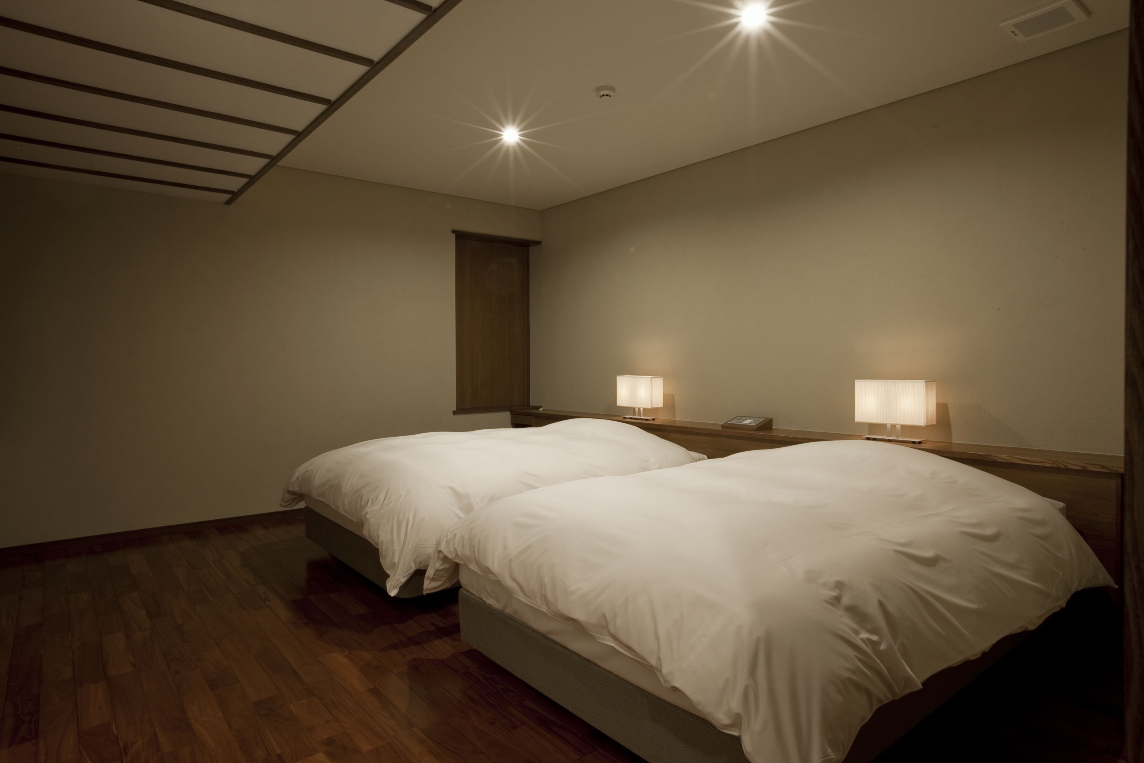A hotel room with two single beds, wooden flooring, a sloped ceiling with recessed lighting, and a nightstand with a lamp.