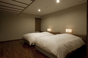 A hotel room with two single beds, wooden flooring, a sloped ceiling with recessed lighting, and a nightstand with a lamp.