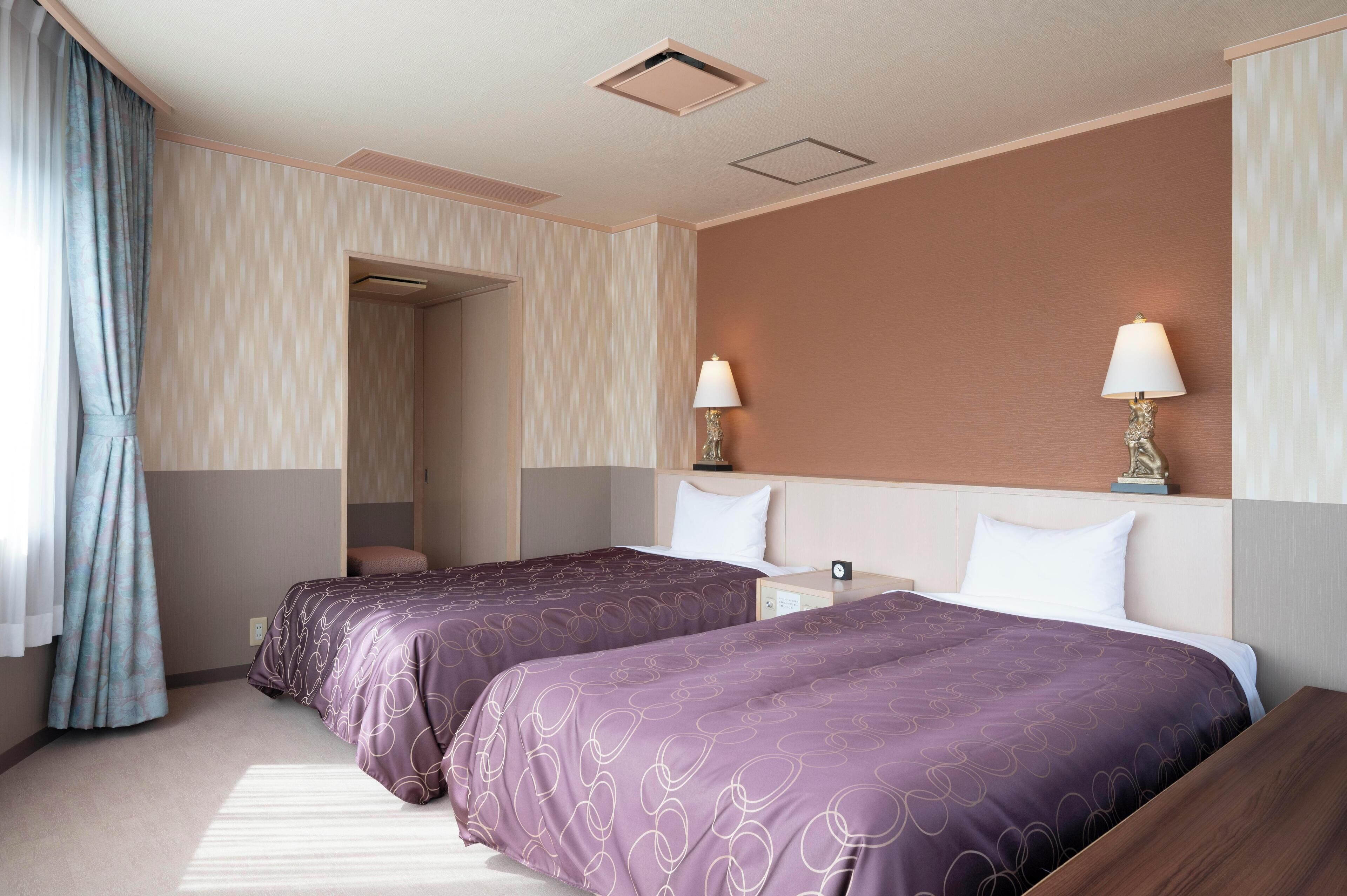 A hotel room with two beds, a nightstand, a lamp, and a window with curtains.