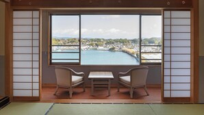 Een traditionele Japanse kamer met tatami vloerbedekking, een lage tafel, twee stoelen en een groot raam met uitzicht op een stad en een waterlichaam.