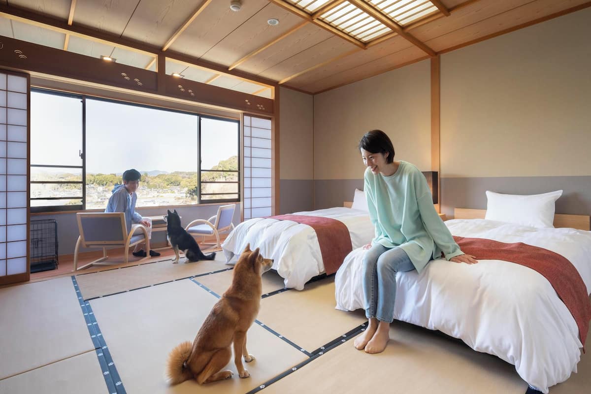 Ett hotellrum med tatamimattor på golvet, en säng med ett rött täcke, ett stort fönster med utsikt över staden och en hund som sitter på golvet.