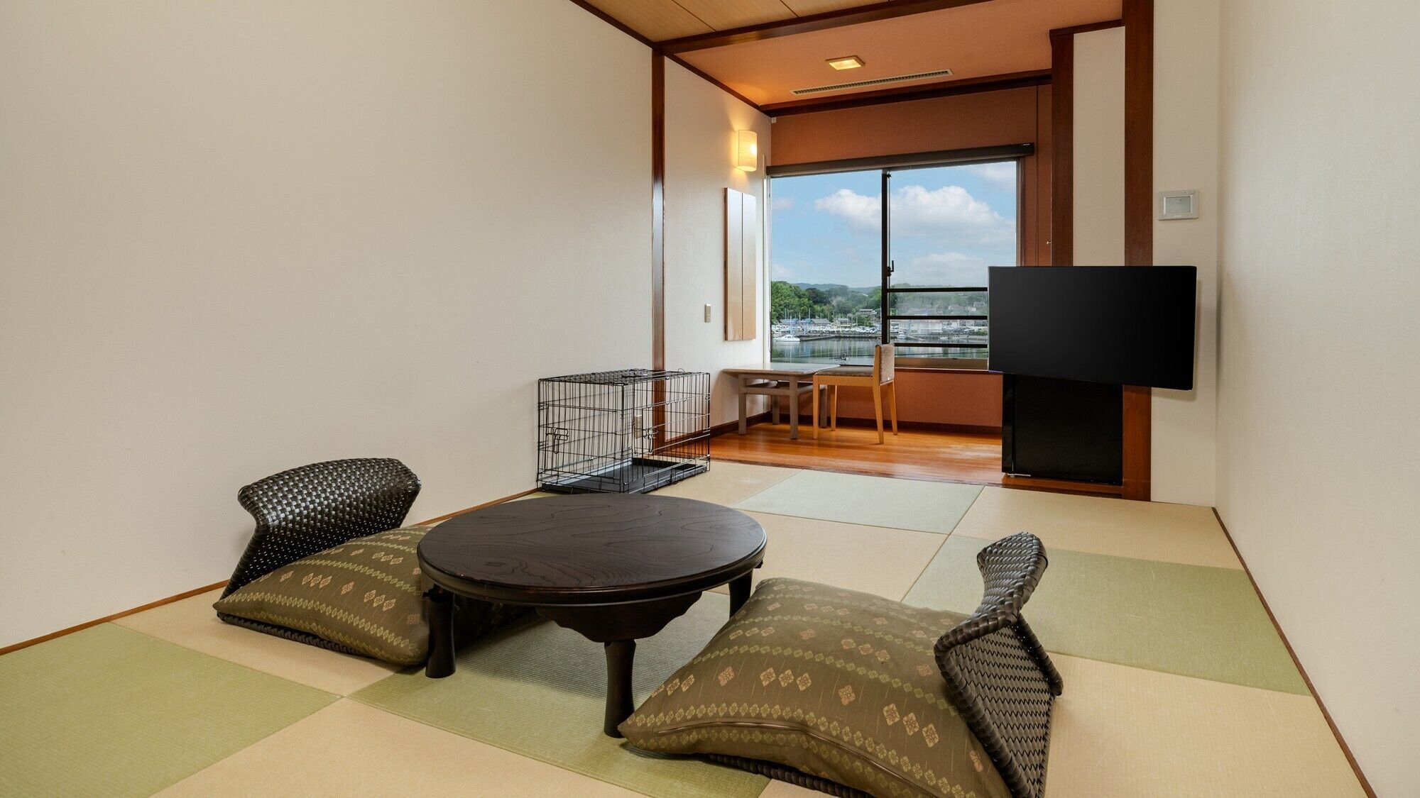 Ein Zimmer mit Tatami-Boden, einem niedrigen Tisch, zwei Kissen, einem Fernseher und Blick auf ein Gewässer durch ein großes Fenster.