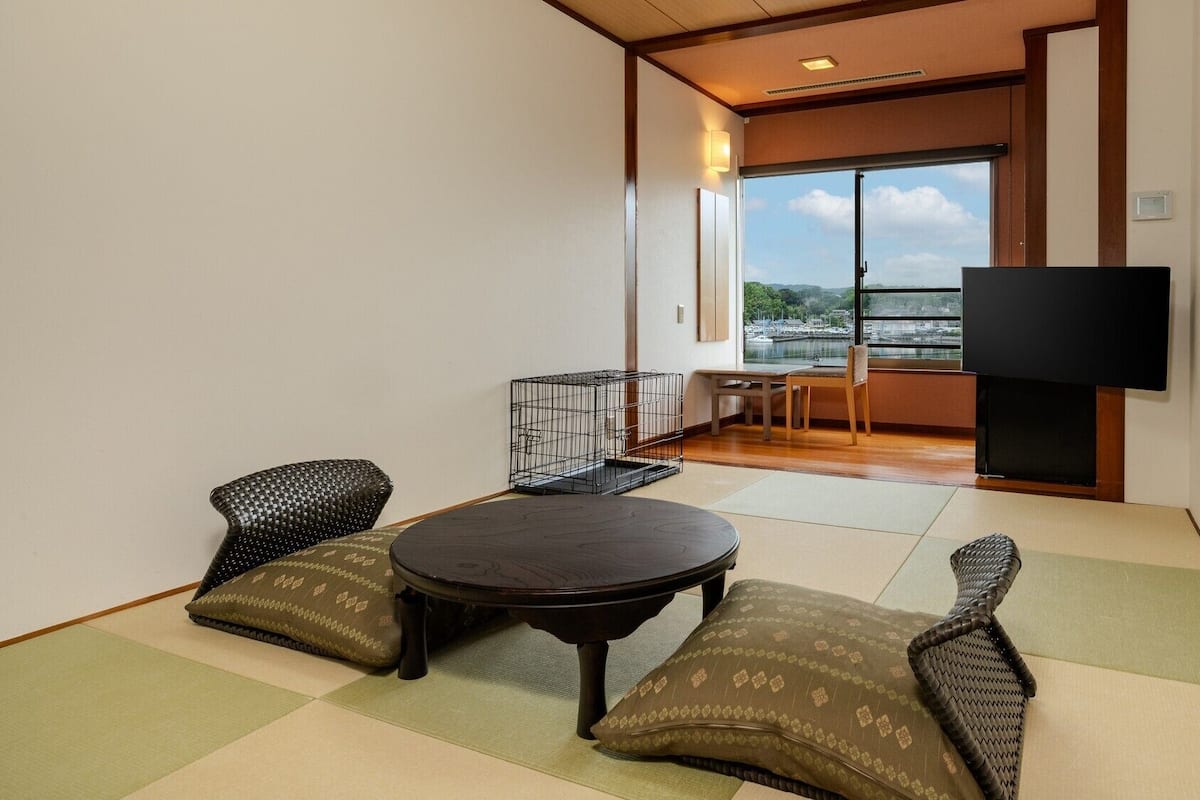 Quarto com piso de tatame, uma mesinha baixa, dois almofadas, uma televisão e vista para um corpo d'água por uma grande janela.