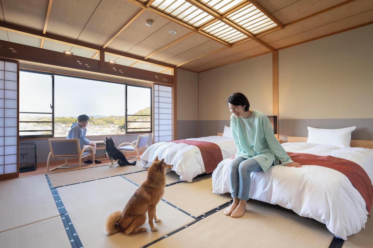 Ett traditionellt japanskt rum med tatamimattor på golvet, en säng med vita sängkläder och utsikt över naturen genom stora fönster.