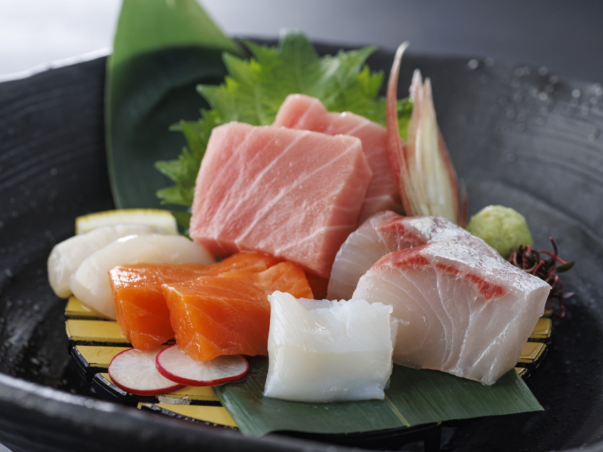 A variety of sushi and sashimi on a black plate with a green leaf underneath.