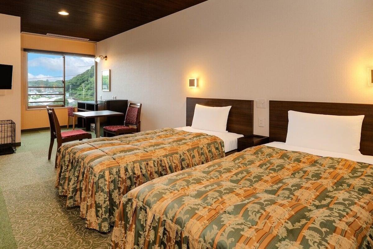 Quarto de hotel com duas camas, uma escrivaninha, uma cadeira, uma televisão e uma janela com vista.