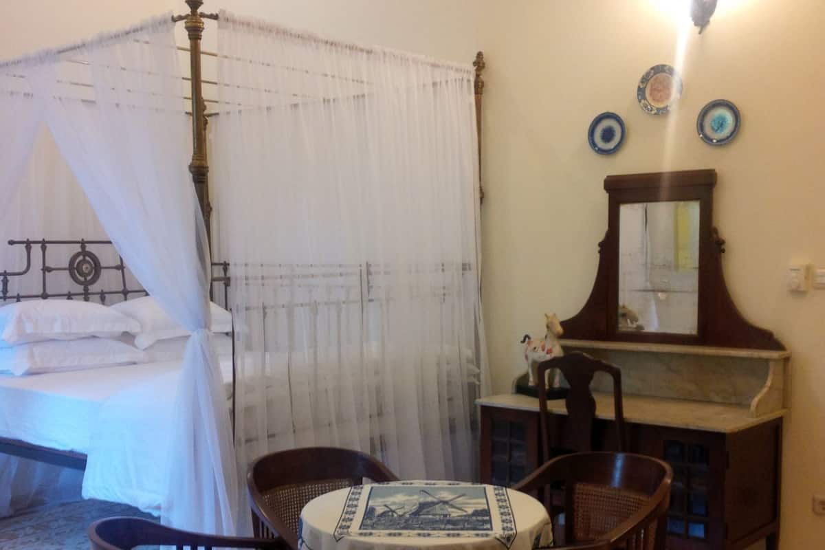 Quarto com cama de dossel, mesa de cabeceira com espelho, pequena mesa redonda com cadeiras e um prato decorativo na parede.