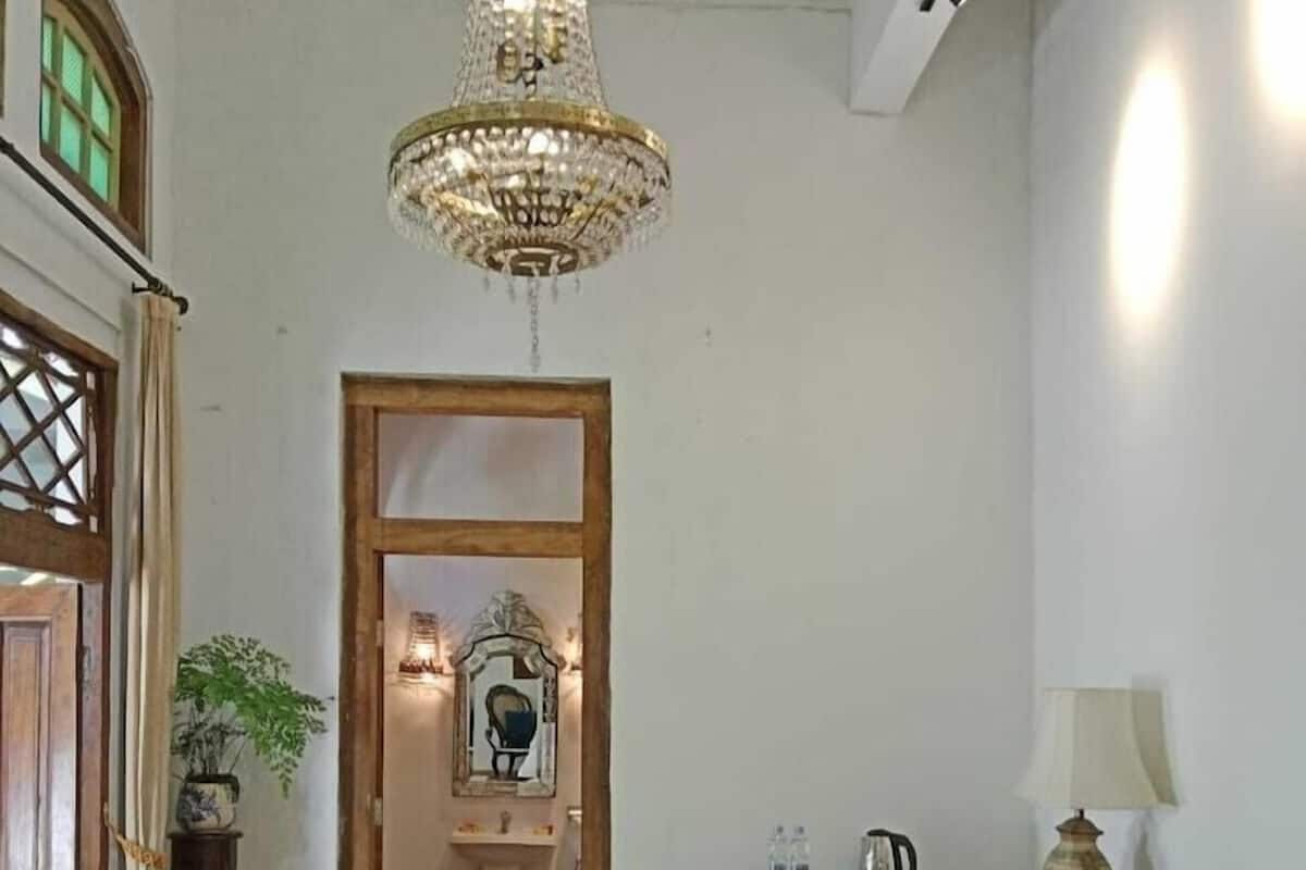Quarto com cama, lustre, espelho, escrivaninha com luminária e janela com cortinas.