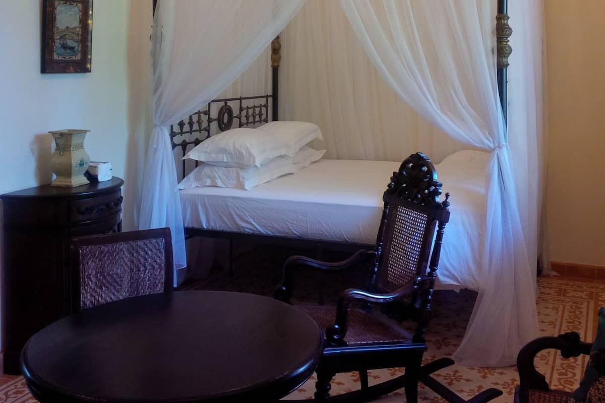 Uma cama com dossel e cortinas, roupa de cama branca, uma pequena mesa com cadeiras e um vaso decorativo em uma mesa de cabeceira.