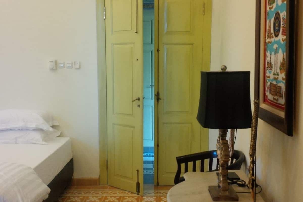 Um quarto com uma cama, uma mesa com abajur e uma porta que dá para outro cômodo.
