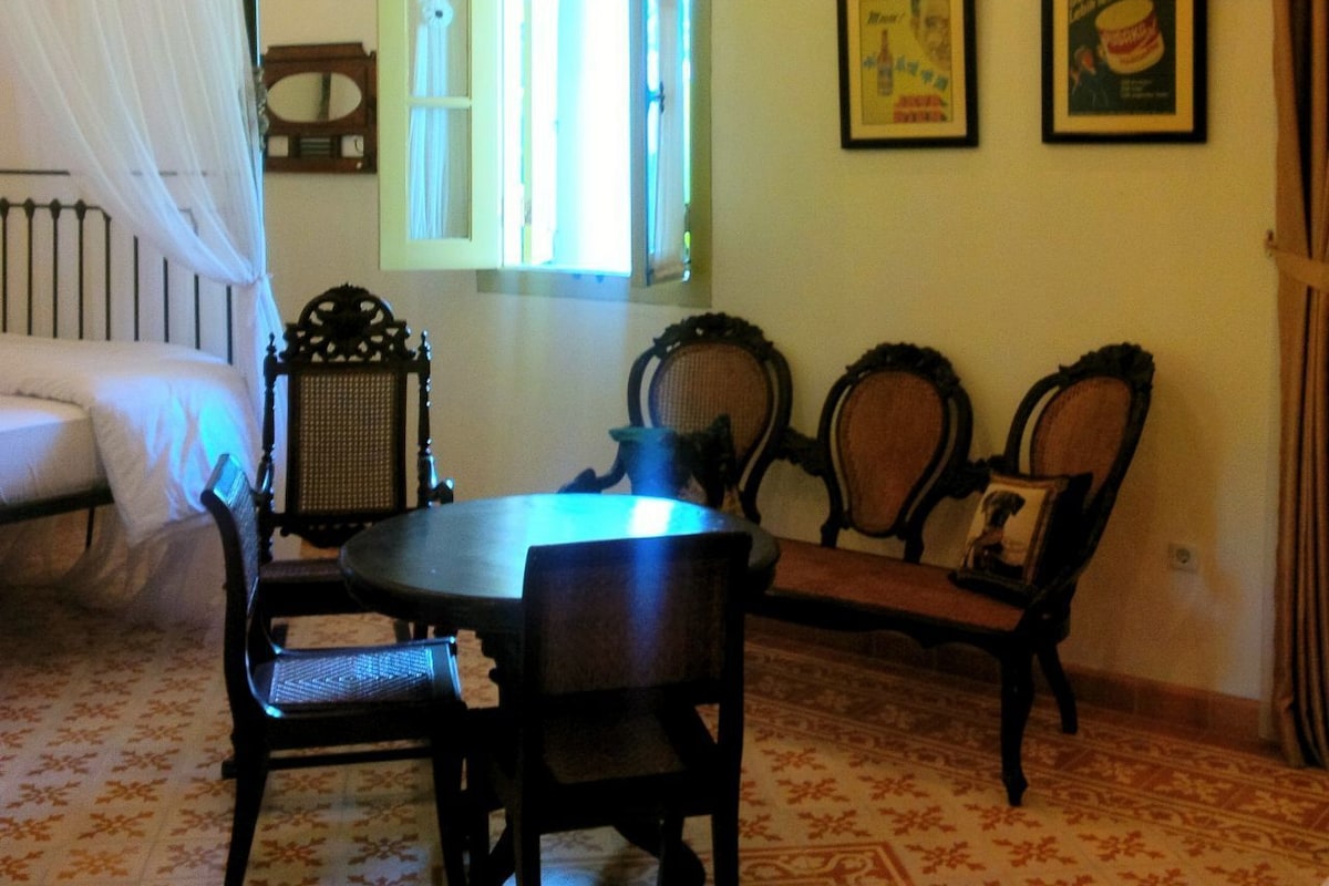 Um quarto com uma cama de dossel, uma mesa redonda com cadeiras, uma janela com cortinas e duas quadros na parede.