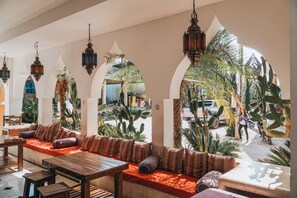 Un'ampia zona salotto con tavolo e panche in legno, circondata da archi e lanterne appese, affacciata su una strada con cactus e palme.