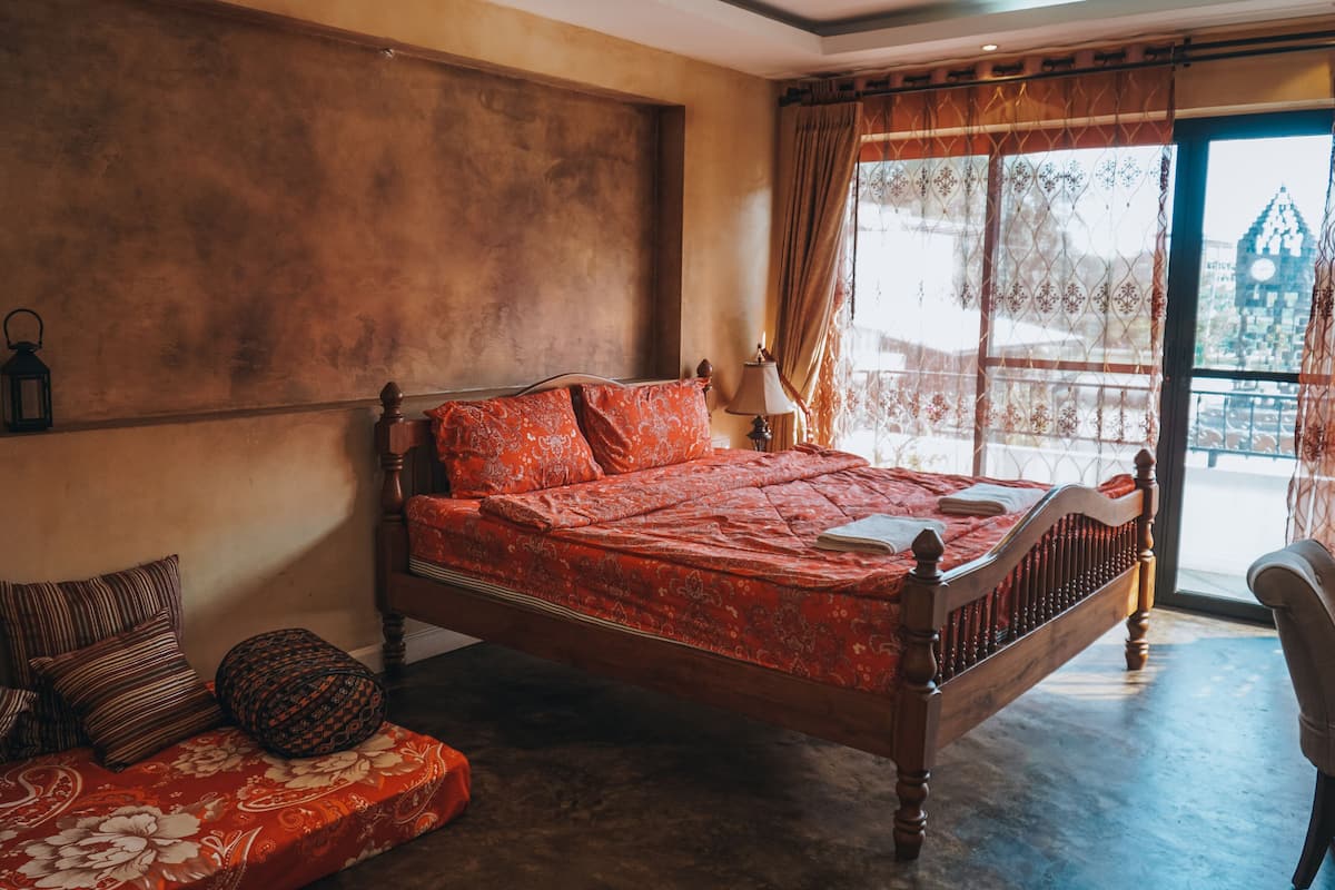 A bedroom with a wooden bed, a bench with cushions, a lamp, and a view of a building through a window.
