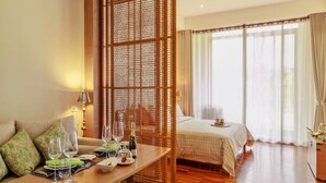 A hotel room with a wooden floor, a bed with a white bedspread, a dining table with glasses and a bottle, and a large window with curtains. - Villa Meesuk Residence (San Sai)