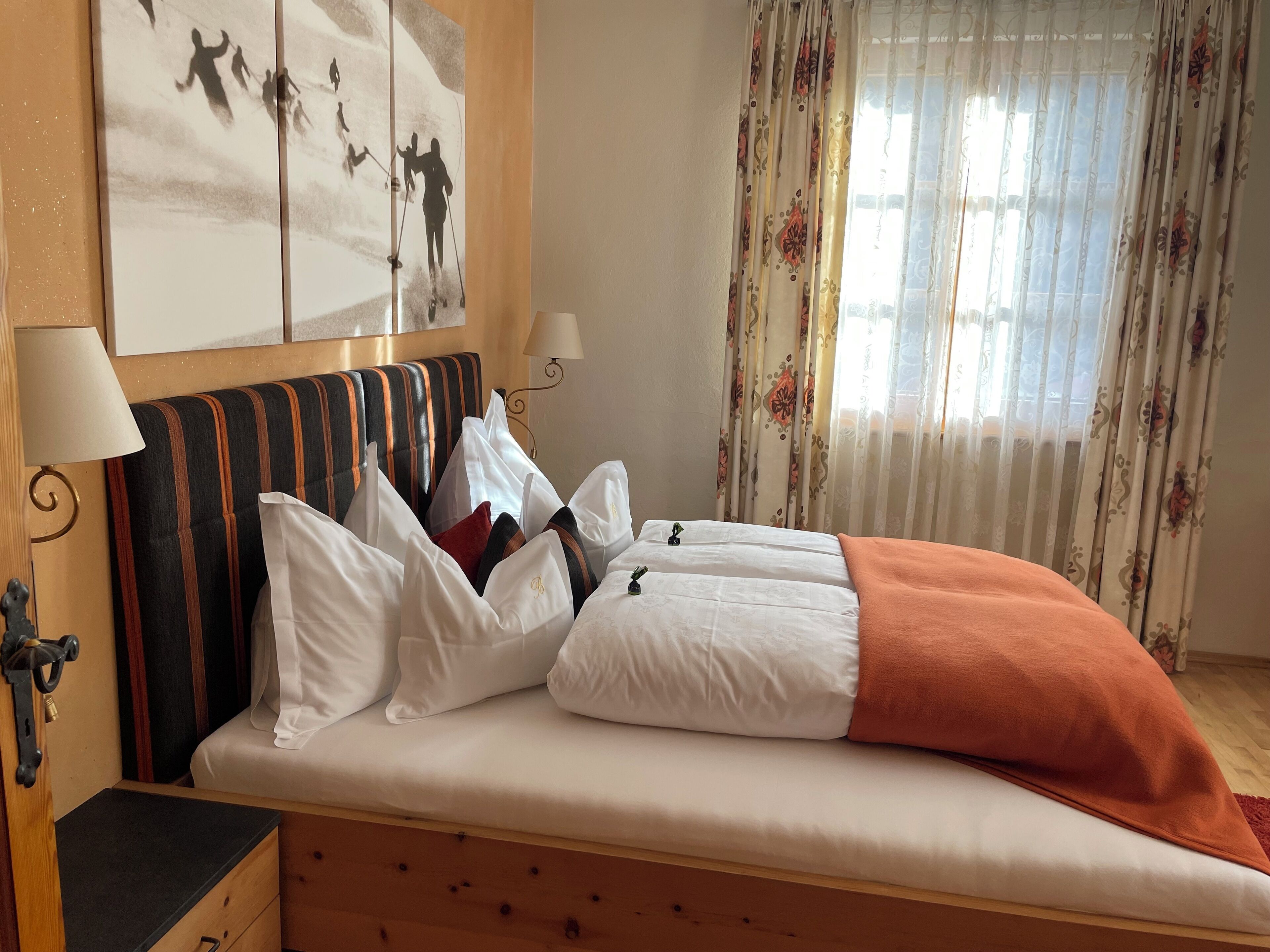 A hotel room with a bed, pillows, a headboard, a lamp, a wall art, and curtains.