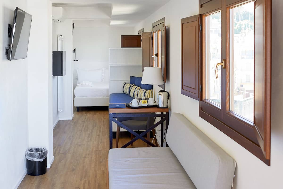 A compact hotel room with a bed, a small table with a lamp, a chair, and a window with wooden shutters.