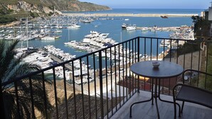 Un balcon avec une table et des chaises donnant sur une marina, où l’on peut admirer des bateaux et une plage.
