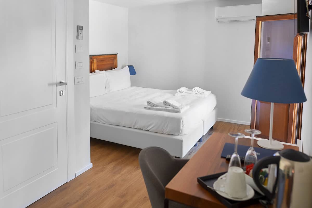 A hotel room with a bed, bedside table, lamp, and a desk with a kettle and glasses.