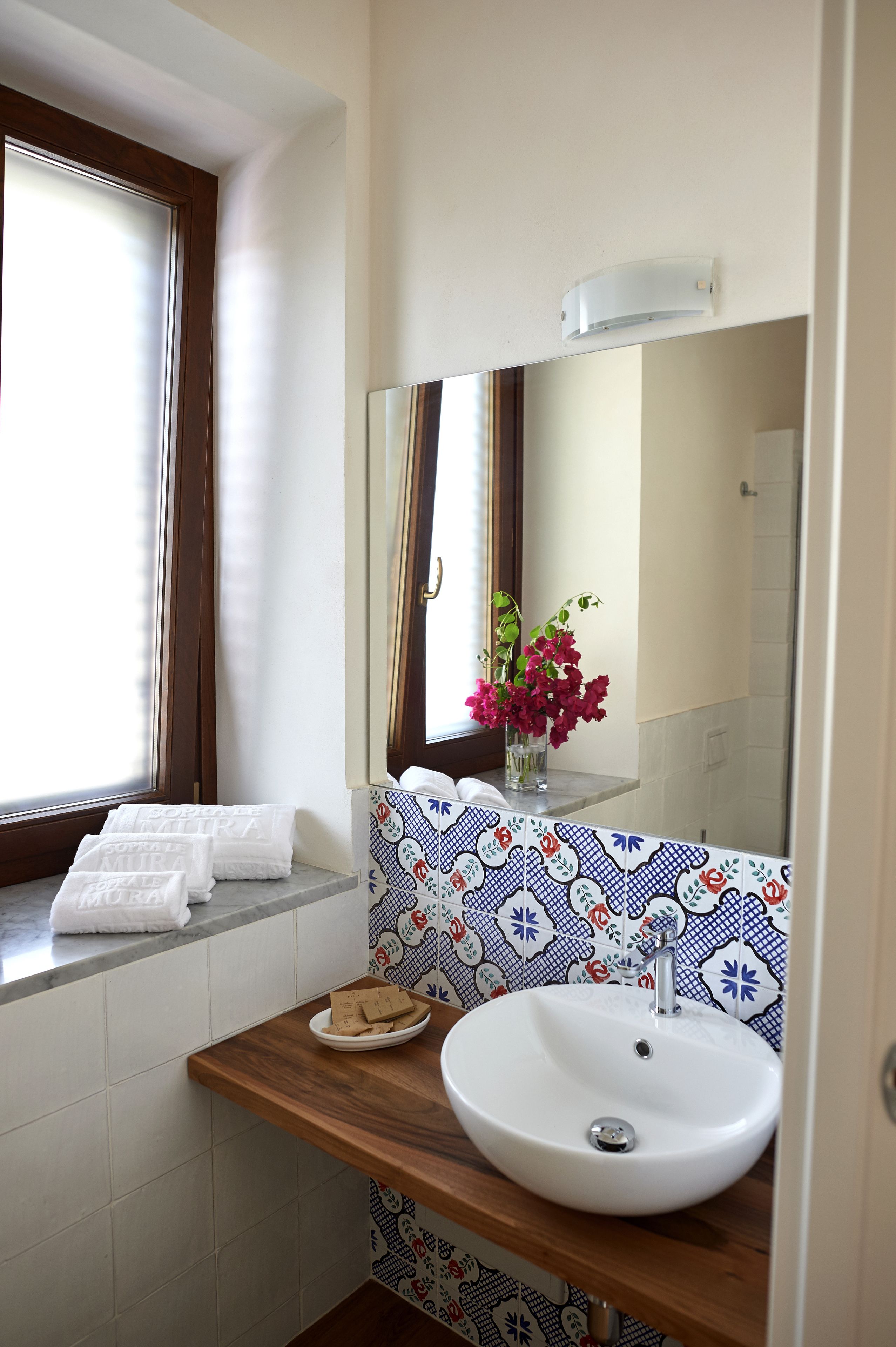 Une salle de bain avec un lavabo blanc, un miroir, une fenêtre avec des rideaux et un vase rempli de fleurs.