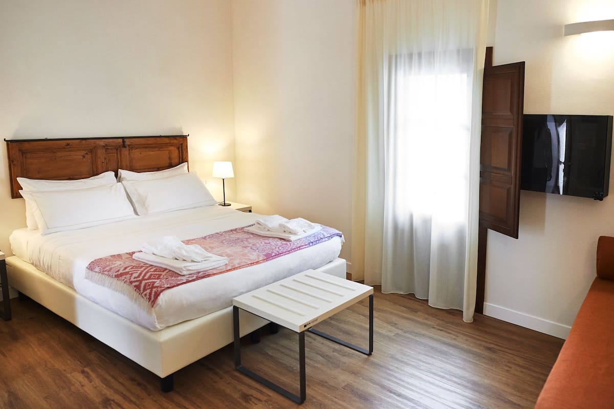 A hotel room with a bed, a bedside table, a TV mounted on the wall, and a window with curtains.