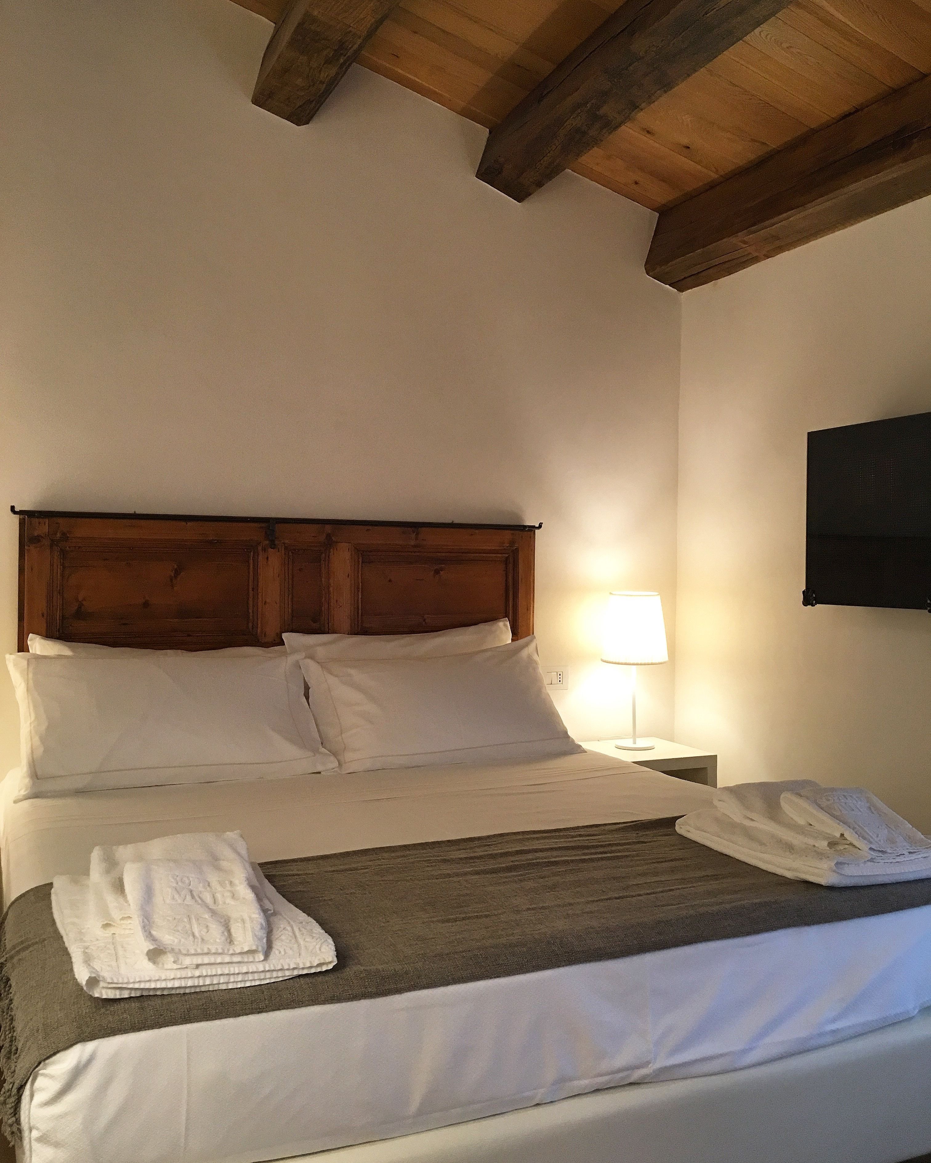 Une chambre d’hôtel avec une tête de lit en bois, un lit recouvert de draps blancs, une lampe de chevet et une serviette pliée posée sur le lit.