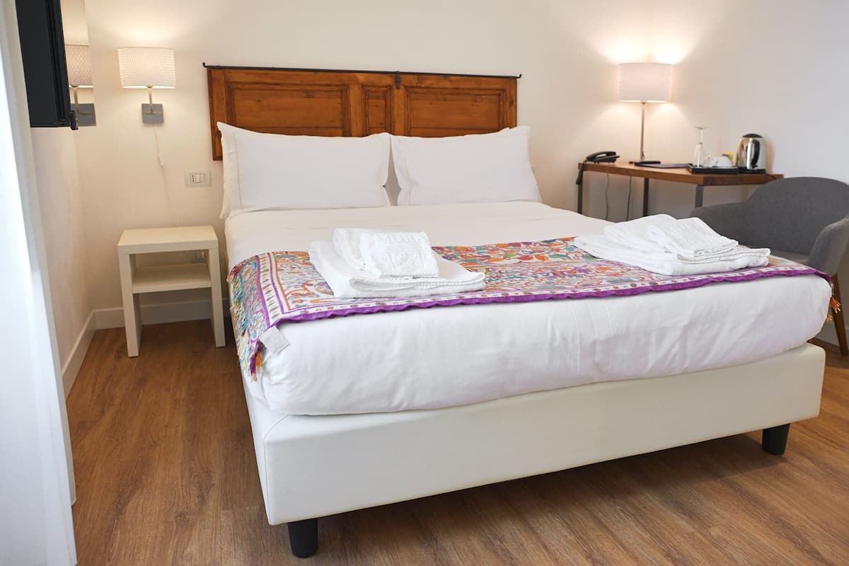 A neatly made bed with white linens and a colorful quilt, a bedside table with a lamp, and a chair in a hotel room with wooden flooring.