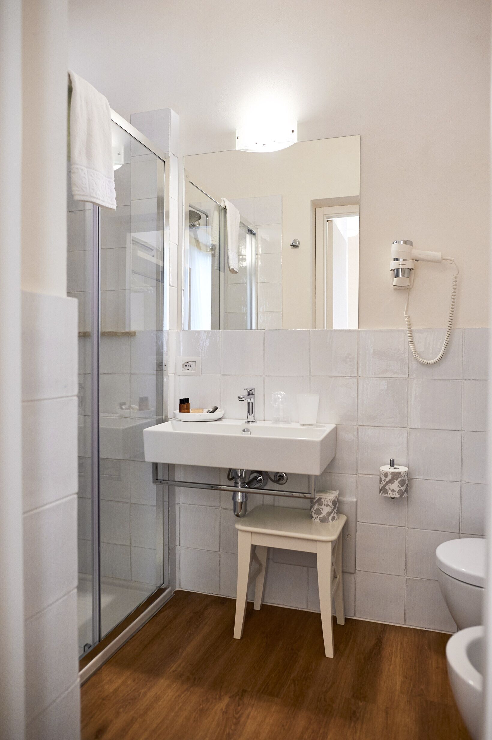 Un baño con cabina de ducha de vidrio, un lavabo blanco con un espejo encima, un taburete pequeño y un inodoro.