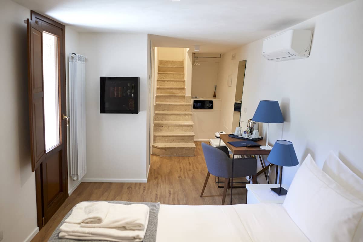 A hotel room with a bed, a desk with a chair, a lamp, and a view of the bathroom and staircase.