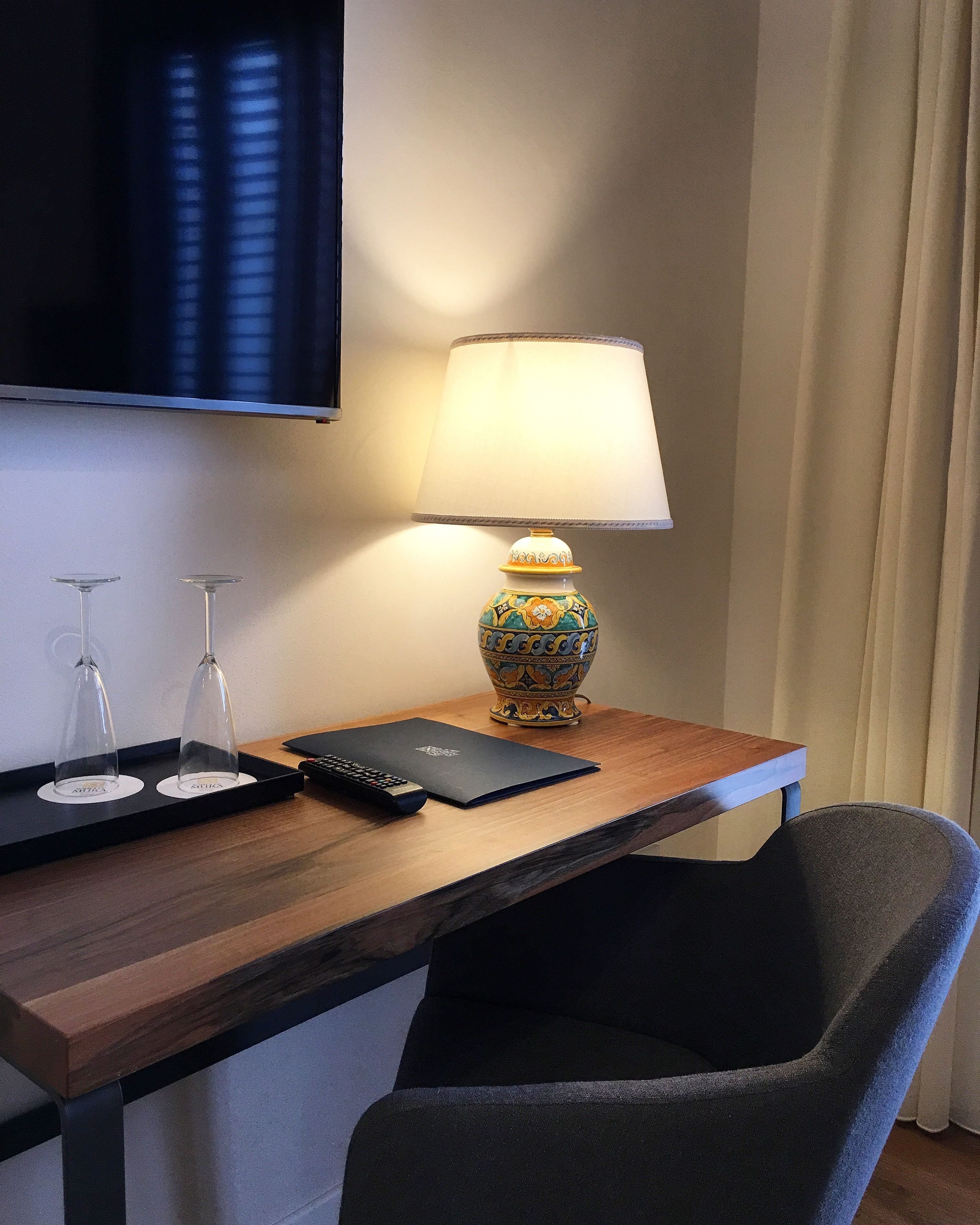 A hotel room with a wooden desk, a lamp, two wine glasses, a remote control, and a laptop.