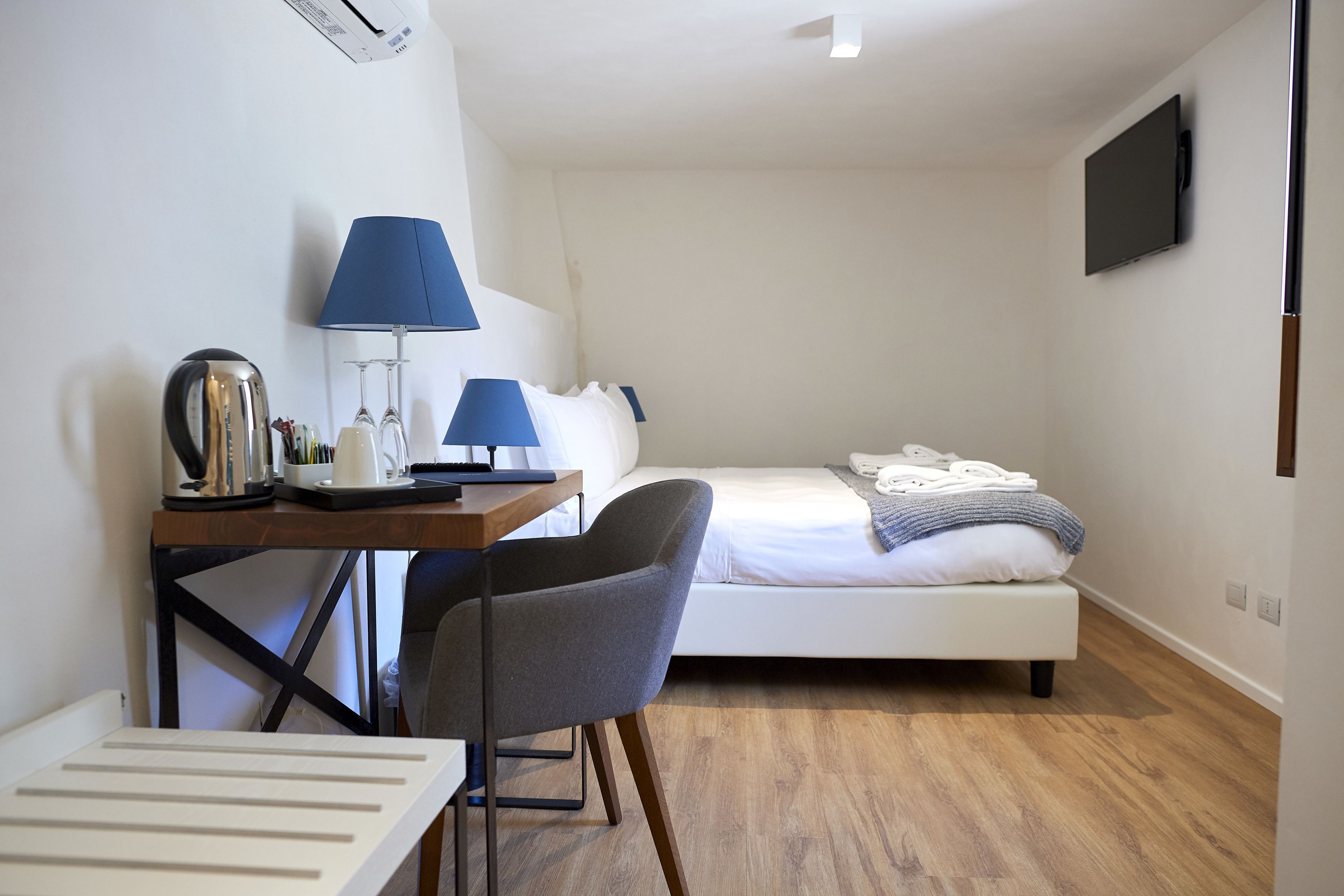 Une petite chambre d’hôtel moderne, équipée d’un lit, d’un bureau avec une chaise, d’une lampe et d’un téléviseur à écran plat fixé au mur.