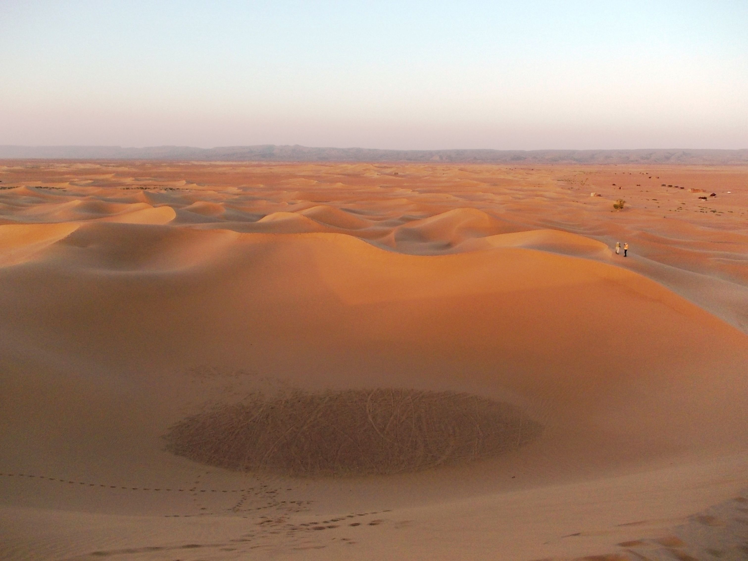 A vast desert landscape with undulating sand dunes and a small group of people in the distance.