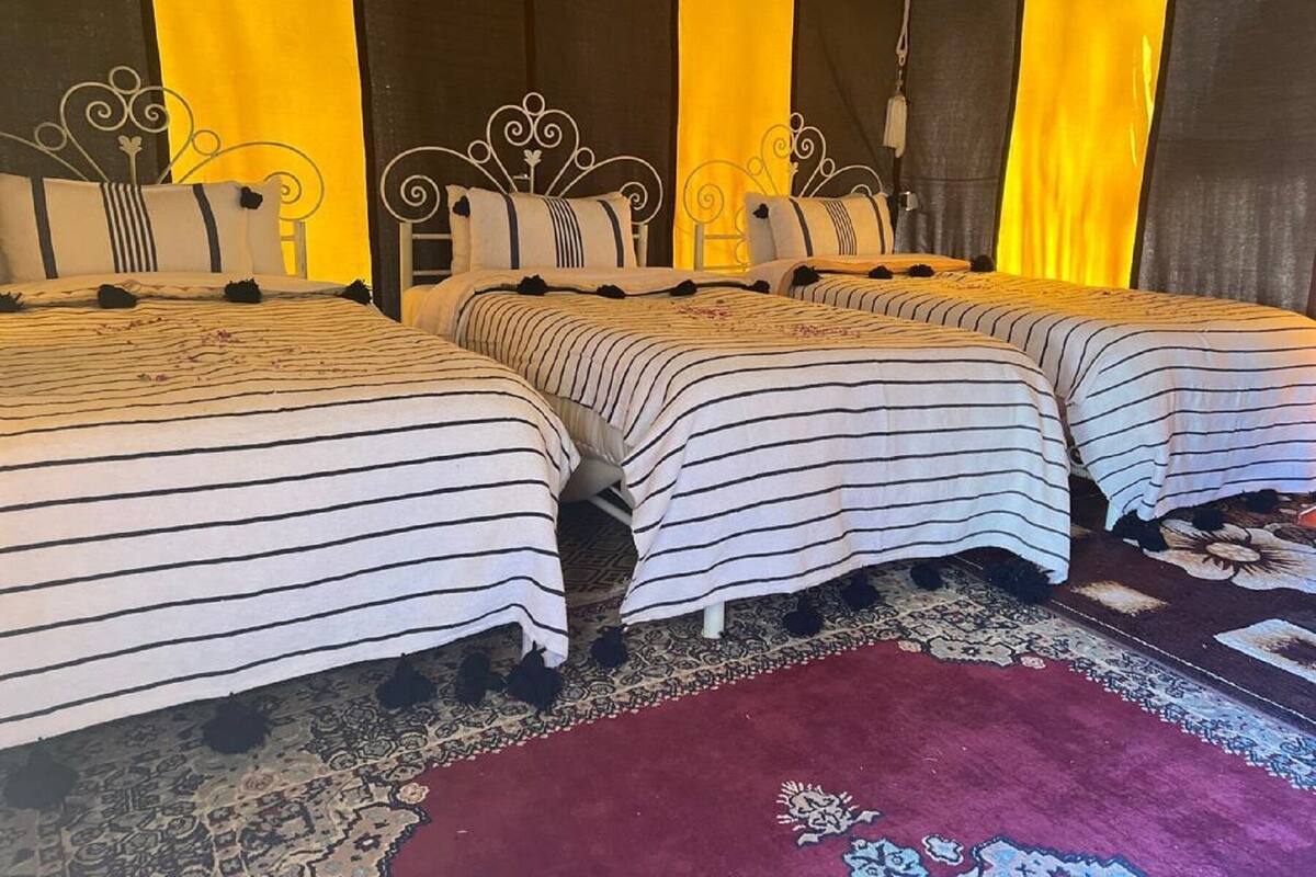 Ein Hotelzimmer mit drei Betten, jedes mit gestreifter Bettwäsche. Der Raum verfügt über einen gemusterten Teppich und eine rote Fußmatte.