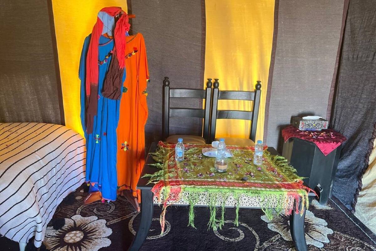 Ein Zimmer mit einem Bett, einem Tisch mit Tischdecke, einem Stuhl und einer mit farbenfroher Kleidung ausgestatteten Mannequin.