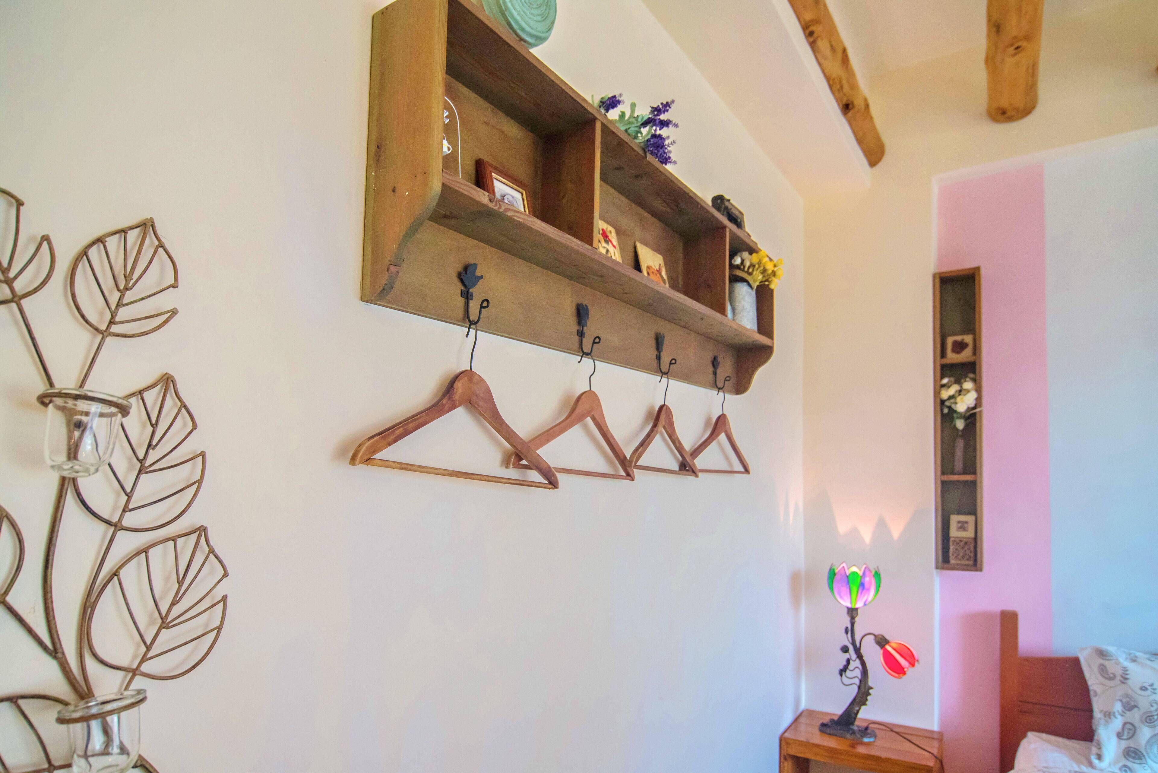 A room with a wooden shelf above a coat rack, a bedside table with a lamp, and a wall-mounted plant.