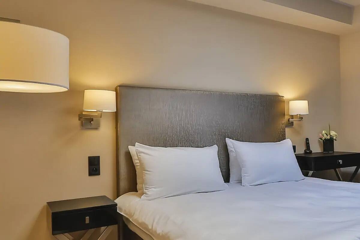A hotel room with a large bed, two bedside lamps, a small table with a flower vase, and a wall-mounted light fixture.