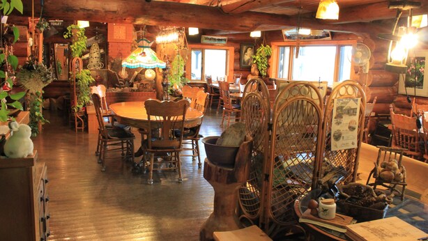 A rustic wooden interior with a round dining table surrounded by chairs, a wooden bench with a basket, and various plants and decorations. - Woody Life (Kamifurano)